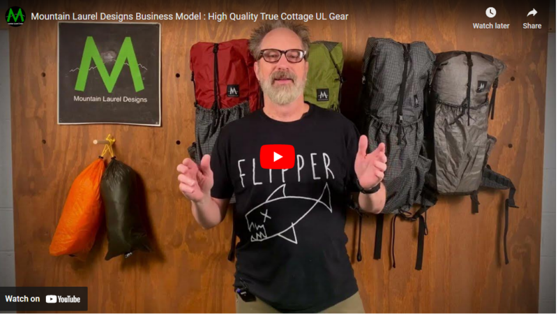 A screenshot of A YouTube video. Ron is standing in the forground, with backpacks hanging on a wood wall behind him.